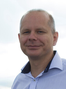 Porträt von Joerg Schmalian mit hellem Hemd vor wolkigem Himmel.